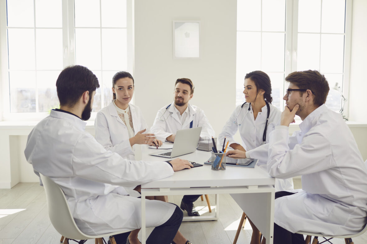 Group of practicing doctors in a meeting discuss the diagnosis of a patient standing in a clinic office.